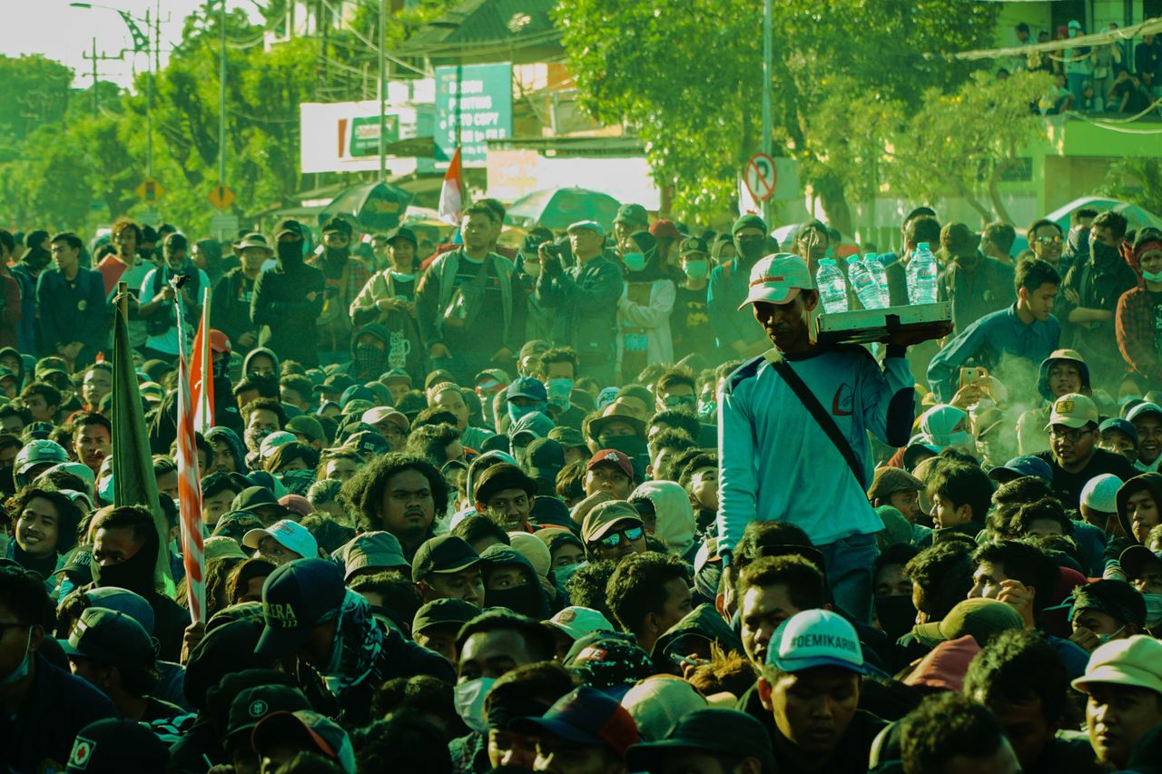 Large crowd of people gathered outdoors with a vendor selling bottled water, creating a lively street scene.